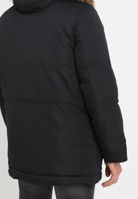 Svart vadderad jacka med huva i fuskpäls och elastiska ärmslut. Har en slät yta och sydda detaljer på baksidan och ärmarna.