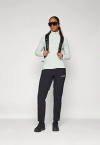 Ženska v sončnih očalih, svetlo sivi zip-up jopič, črne pohodne hlače, pohodniški čevlji in črn nahrbtnik, katere naramnice so vidne na ramenih.