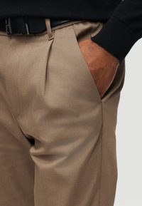 Main dans la poche d'un pantalon beige porté avec une ceinture noire et un haut à manches longues noir, montrant la texture du tissu et les détails de la couture.