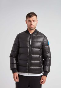 Giacca a vento nera con chiusura a zip, forma squadrata e polsini a coste. Presenta un logo sulla zip e un accento blu sulla manica.