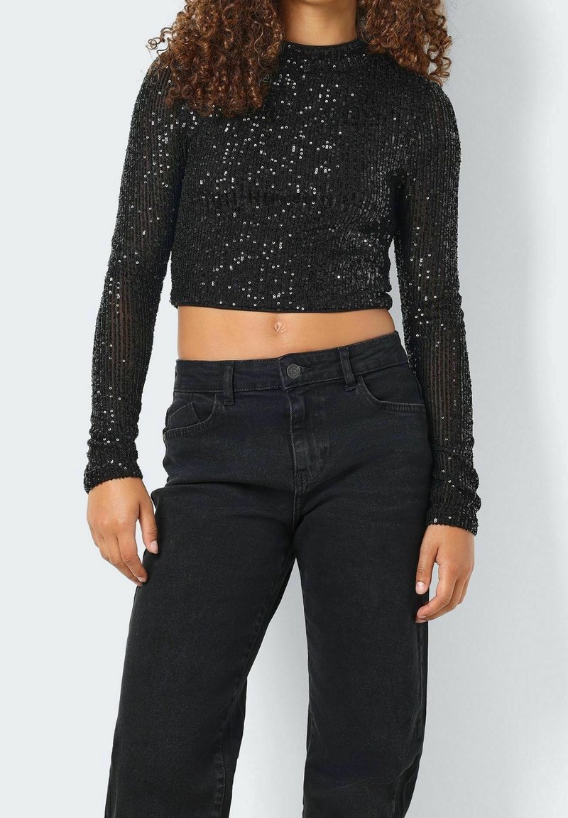 Femme portant un crop top noir à manches longues pailleté et un jean noir taille haute, debout avec une main détendue le long du corps.