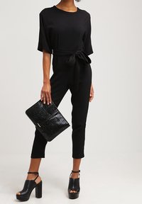 Tuta nera con maniche corte e vita legata. Abbinata a una pochette nera con texture a coccodrillo e sandali neri con tacco grosso.