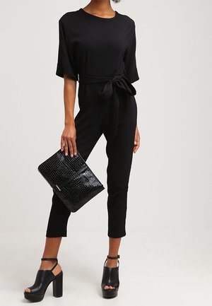 Czarny kombinezon z krótkim rękawem i wiązanym w pasie. Połączony z czarną torebką typu kopertówka w krokodylą fakturę i masywnymi czarnymi sandałami na obcasie.