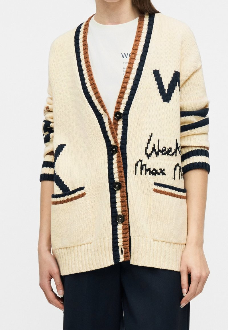 Gilet en tricot crème avec bordures noires et marron, boutons noirs, grandes lettres noires, poches avant, porté sur une chemise blanche et un pantalon foncé.