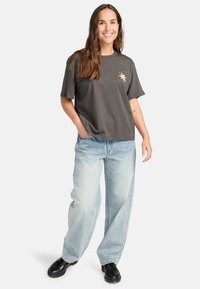Grå bomulds T-shirt med korte ærmer og et grafisk design på fronten, parret med lyseblå baggy jeans og sorte sko.