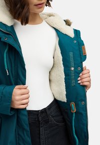 Parka teal con fodera in pelliccia sintetica bianca e hardware argentato. Presenta una chiusura con zip, cordini regolabili e un accento tasca tonale.