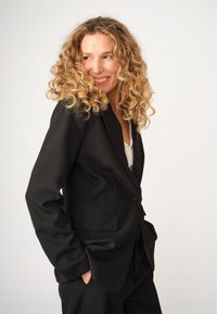 Schwarze Blazer mit taillierter Passform, einreihiger Knopfschluss und Fronttaschen. Hergestellt aus strukturiertem Stoff, mit breiten Revers und lockerer Passform.