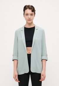 Helles grünes, offen gestaltetes Blazer mit drei Viertelärmel, zwei Taschen und einem strukturierten Stoff. Verfügt über goldene Knopfakzente an den Ärmeln.