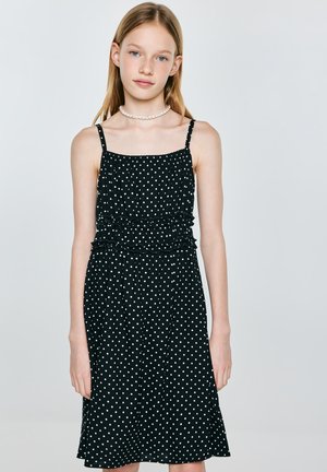 Młoda dziewczyna ubrana w czarną, bezrękawną sukienkę w białe kropki oraz perłowy naszyjnik, stojąca na jednolitym jasnym tle.