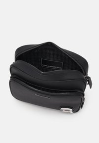 Borsa nera con zip e due scomparti, realizzata in materiale testurizzato. L'interno presenta un motivo houndstooth e un'etichetta con logo. Finiture in metallo argentato.
