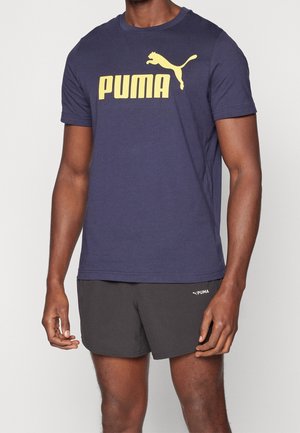 Hombre con una camiseta azul marino Puma con logo amarillo y pantalones cortos negros Puma, de pie frente a un fondo claro y liso.