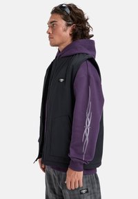 Gilet senza maniche nero sopra un hoodie viola; presenta una zip frontale, grandi tasche e una piccola patch con logo sul gilet. Visibili pantaloni a scacchi casual.
