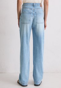 Osoba w jasnoniebieskich, szerokich dżinsach z tylnymi kieszeniami, stojąca przy gładkiej, białej ścianie.