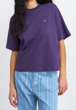 Kobieta ubrana w luźny fioletowy t-shirt z krótkim rękawem i małym logo oraz jasnoniebieskie, paskowane jeansy z wysokim stanem na jednolitym tle.