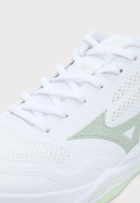 Gros plan d'une chaussure de sport blanche avec un logo vert clair, un tissu en mesh et des lacets blancs sur un fond neutre.