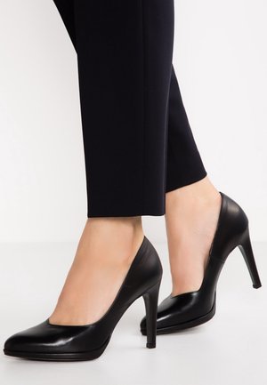 Vrouw die zwarte leren pumps met hoge hakken en puntige neuzen draagt en korte zwarte broek tegen een witte achtergrond.