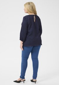 Blusa navy con tessuto testurizzato, maniche lunghe a sbuffo, dettaglio ovale sulla parte posteriore, abbinata a jeans skinny blu e mule neri con tacco.