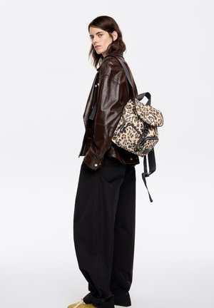 Kobieta w ciemnobrązowej skórzanej kurtce i luźnych czarnych spodniach, niosąca na jednym ramieniu beżowy plecak w cętki lamparta.