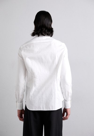 Witte longsleeve shirt met een gestructureerd patroon van herhaalde branding, voorzien van een afgeronde zoom en knopenmanchetten, gecombineerd met donkere broeken.