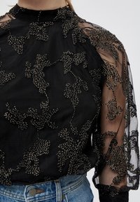 Blouse noire avec des manches longues transparentes. Présente des perles en argent intricatement disposées en motifs floraux. Col montant et tissu texturé.