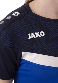 Maglietta sportiva blu e blu navy scuro con motivo testurizzato, dettagli bianchi e logo "JAKO" sul petto, presenta un logo rotondo sulla spalla.