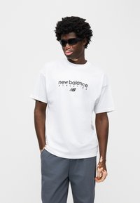 Λευκό κοντομάνικο μπλουζάκι με το λογότυπο «new balance» σε μαύρο, συνδυασμένο με γκρι παντελόνια. Τα αξεσουάρ περιλαμβάνουν σκούρα γυαλιά ηλίου.