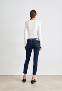 Camiseta blanca de manga larga combinada con vaqueros ajustados de color azul oscuro, con una etiqueta de marca visible en la parte trasera de la cintura y un diseño sencillo. Zapatos de tacón negros.