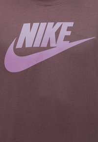 Το λογότυπο της Nike με έντονα γράμματα και σύμβολο swoosh σε ανοιχτό μωβ χρώμα πάνω σε φόντο από ύφασμα σε απαλό καφέ χρώμα.