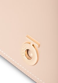 Pfirsichfarbene Tasche aus weichem Leder mit goldfarbener runder Hardware mit Logo und einem rechteckigen Akzent darüber. Weiße Nähte.