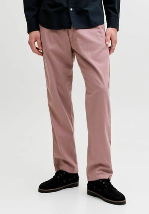 Mand iført pink bukser med lige ben, sorte snøresko og en sort skjorte med knapper, stående mod en ensartet lys baggrund.