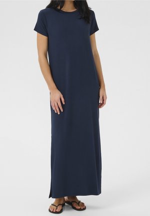 Kvinde iført en mørkeblå, kortærmet, ankel-lang afslappet kjole med sideslids og sorte thongsandaler foran en ensfarvet baggrund.