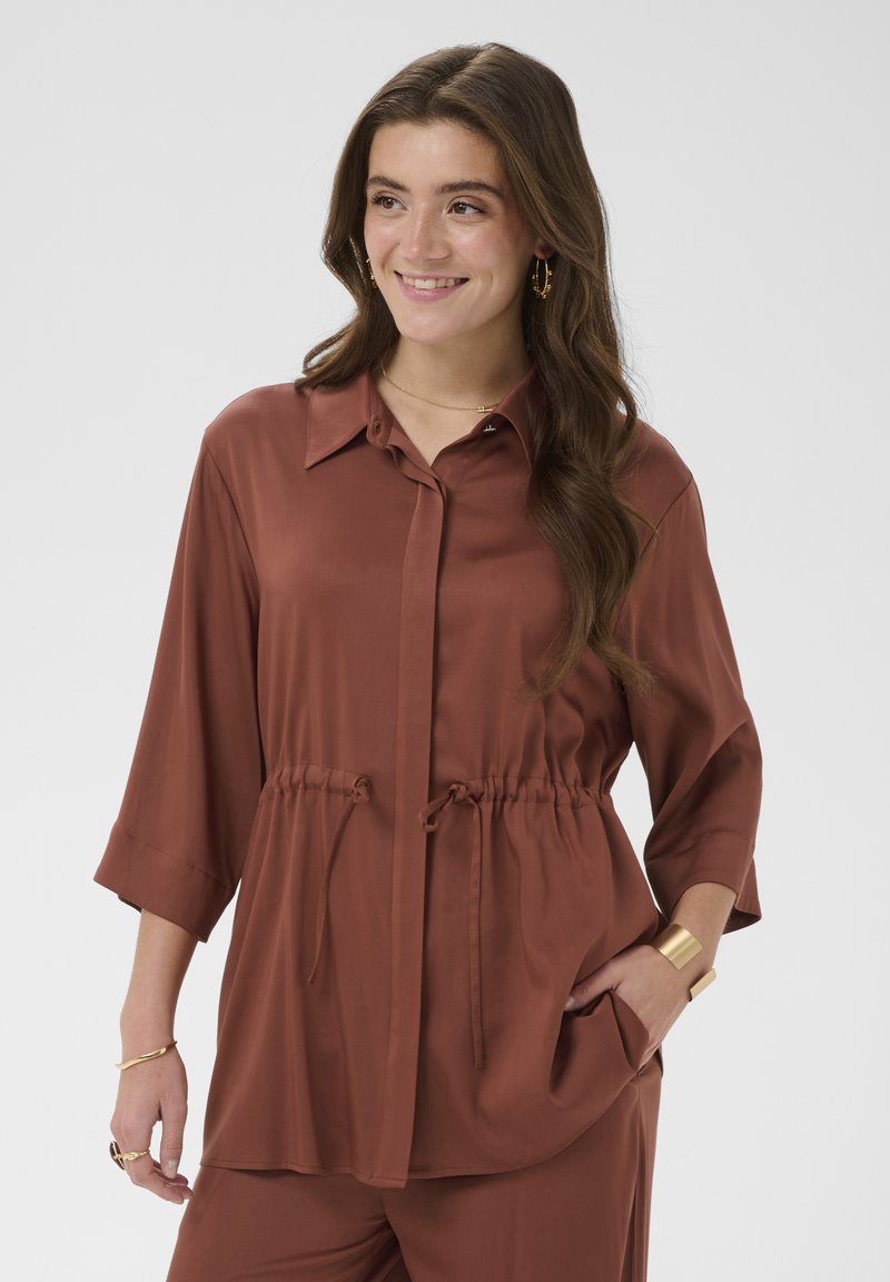 Femme portant un haut boutonné couleur rouille avec lien à la taille et pantalon assorti, souriante, la main dans la poche sur fond uni.