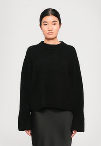 Una donna con capelli neri lisci raccolti indossa un maglione nero morbido e largo e una gonna nera liscia, il tutto su uno sfondo grigio semplice.