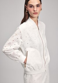 Giacca bomber bianca con un motivo a occhio di pernice testurizzato, polsini a costine e chiusura con zip. Presenta una tasca laterale singola con hardware dorato.
