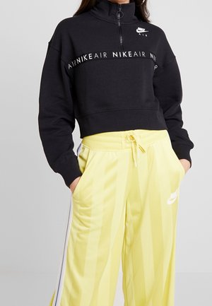 Czarna krótka bluza Nike Air z półzamkiem, w komplecie z żółtymi dresowymi spodniami Nike z wysokim stanem i białymi paskami po bokach.