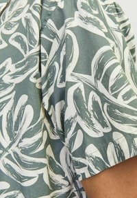 Camicia a maniche corte in tessuto verde con motivo di foglie bianche. Il design presenta una texture morbida e una vestibilità rilassata. Cuciture visibili di notevole fattura.