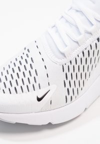 Detailní záběr na bílou tenisku Nike s černými ventilačními otvory a malým černým znakem Nike poblíž podrážky.