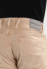 Beige denim jeans with a classic five-pocket design. Features a gray logo patch labeled "REPLAYJEANS SINCE 1981." Textured fabric surface.