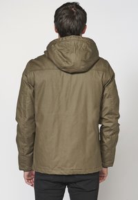 Mężczyzna w oliwkowym długim płaszczu z kapturem widziany od tyłu, w połączeniu z czarnymi spodniami, na jednolitym białym tle.