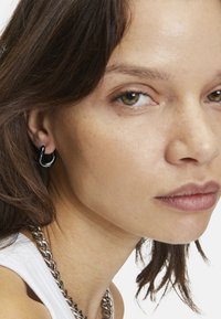 Zwarte hoepeloorbellen met een glanzende afwerking, gedragen in het oor, vergezeld van een dikke zilveren kettingketting, tegen een neutrale achtergrond.