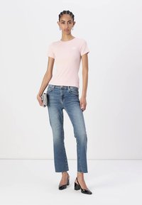 T-shirt ajusté rose clair avec logo blanc, associé à un jean droit bleu. Le modèle porte des chaussures à talons noirs et tient une pochette argentée.