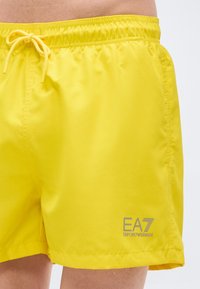 Κίτρινο μαγιό σορτς από ελαφρύ ύφασμα, με ελαστική μέση και κορδόνι ρύθμισης, καθώς και γκρι εκτύπωση λογότυπου στην πλευρά.