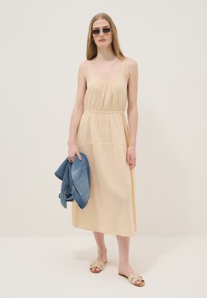 Vrouw draagt een beige midi-jurk en sandalen, houdt een blauw spijkerjasje vast, met steil haar en een zonnebril tegen een effen achtergrond.