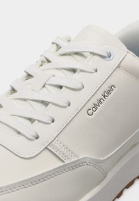 Κοντινό πλάνο λευκού αθλητικού παπουτσιού Calvin Klein που δείχνει τα κορδόνια, τις ραφές και το λογότυπο στο λείο δερμάτινο πάνω μέρος.