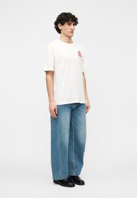 Λευκό βαμβακερό t-shirt με κοντά μανίκια και κόκκινο γραφικό σχέδιο. Συνδυάζεται με ανοιχτό μπλε τζιν παντελόνι φαρδιάς γραμμής και μαύρα παπούτσια.