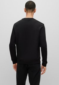 Hombre de espaldas vestido con un sudadera negra de manga larga y pantalones negros sobre un fondo gris claro.