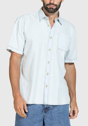Uomo che indossa una camicia azzurro chiaro a maniche corte con taschino sul petto e jeans blu, in piedi davanti a uno sfondo semplice.