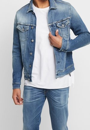 Mann trägt blaue Denimjacke, weißes T-Shirt und blaue Jeans, mit einer Hand passt er die Jackenvorderseite in Brustnähe an.