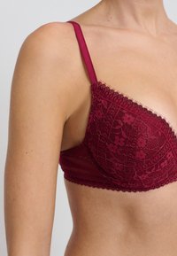 Bralette in pizzo bordeaux con motivo floreale, coppe triangolari, fascia elastica e spalline regolabili. Presenta bordi smerlati e dettagli fini.