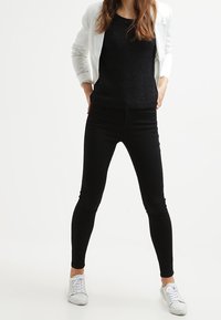 Jeans skinny neri, materiale elasticizzato, design a vita alta; abbinati a un top a maglia nera e a un blazer bianco; sneakers bianche completano l'outfit.